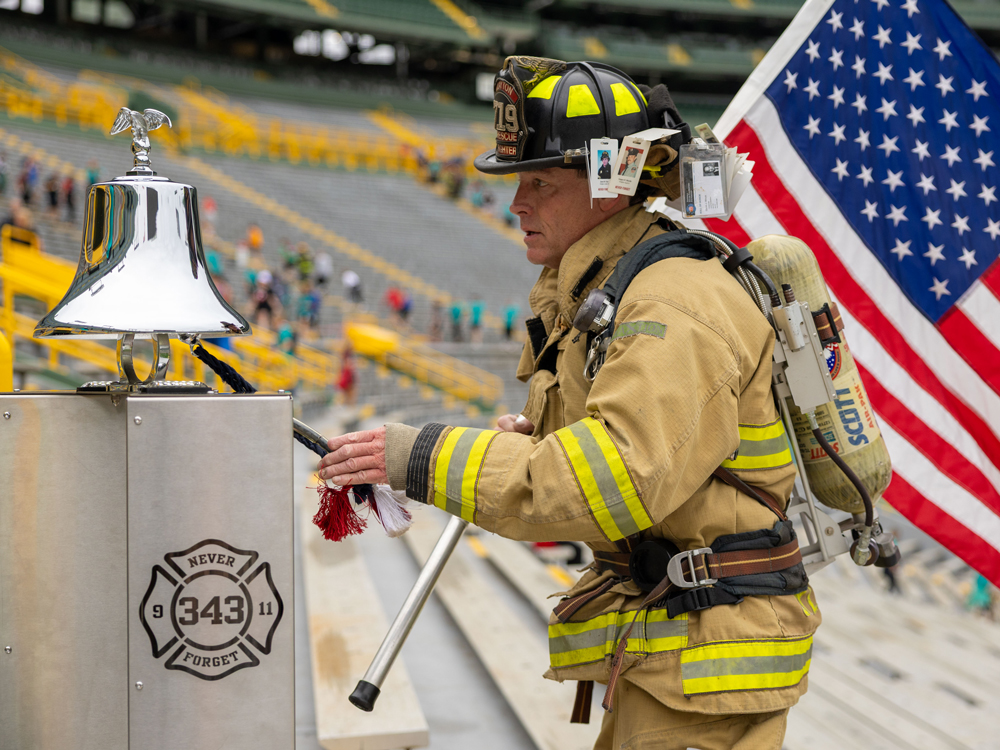 Firefighter holding an American flag and ringing the bell at the 2025 Pierce Manufacturing Stair Climb event at Lambeau Field